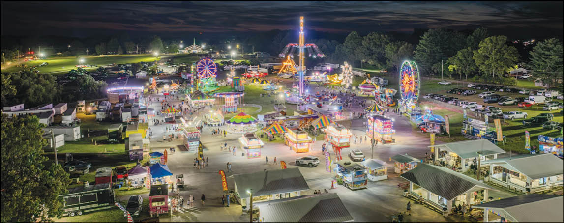A brightly lit nighttime carnival scene with various rides, booths, and people scattered throughout an open area surrounded by trees and parked vehicles. Description generated by AI
