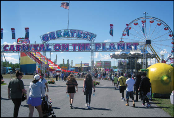 People walk toward a colorful carnival entrance sign that reads “Greatest Carnival on the Planet" with rides and a Ferris wheel visible in the background under a clear blue sky. Description generated by AI