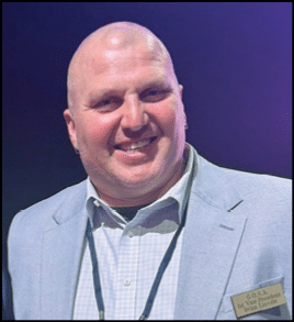 A smiling adult wearing a light gray blazer and checkered shirt with a name tag that reads “G.O.S.A 1st Vice President Brian Linville" against a dark background. Description generated by AI