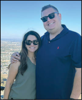 Two adults wearing sunglasses smile and pose closely together outdoors with a cityscape visible in the background. Description generated by AI