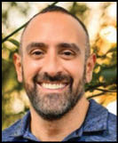 A close-up of the upper part of a person's head with short dark hair and a blurred natural background. Description generated by AI