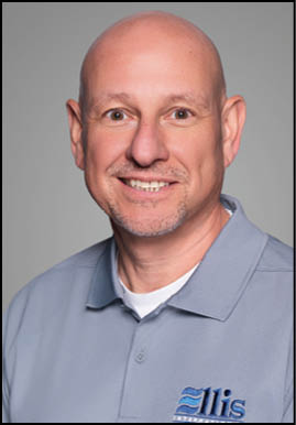 A smiling adult with a bald head wearing a light blue collared shirt with a logo on it is posed against a plain gray background. Description generated by AI