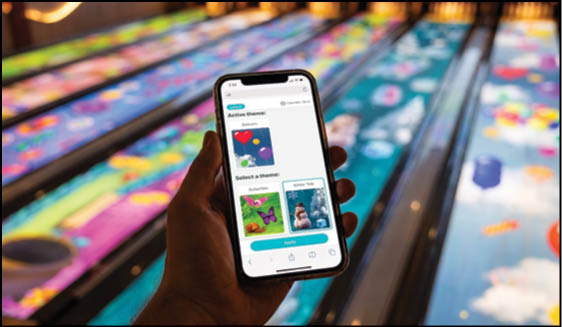 A person is holding a smartphone displaying an app screen with colorful themes while standing near a brightly lit bowling alley lane. Description generated by AI