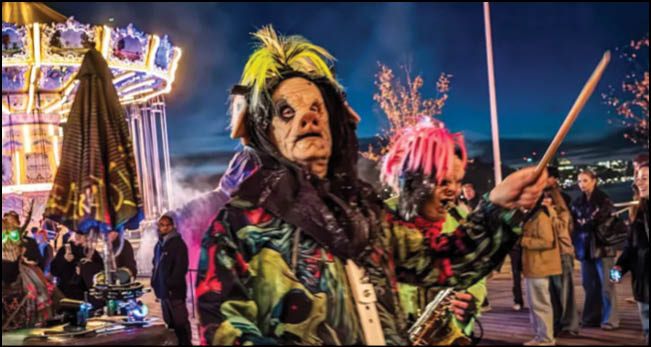People wearing colorful costumes and masks are participating in a lively outdoor event near a brightly lit carousel at night. Description generated by AI