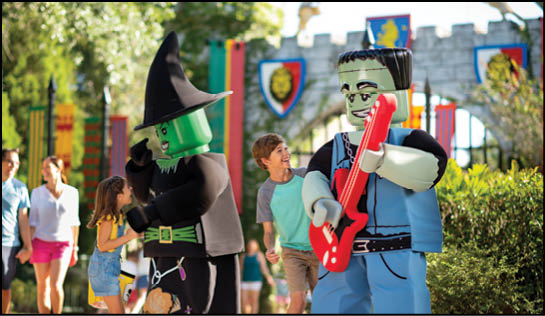 Two people in large Halloween-themed costumes, one dressed as a witch and the other as Frankenstein playing a red guitar, interact with children in a colorful outdoor setting with castle-like decorations. Description generated by AI