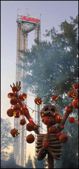 A large skeleton decoration holding strings of glowing jack-o'-lanterns is set outdoors near a tall amusement park ride tower with the word Vallejo visible at the top. Description generated by AI