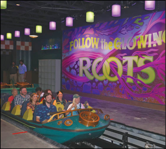 A group of people seated in a green boat ride vehicle inside an indoor attraction with colorful lighting and a large mural that reads “FOLLOW the GLOWING ROOTS." Description generated by AI