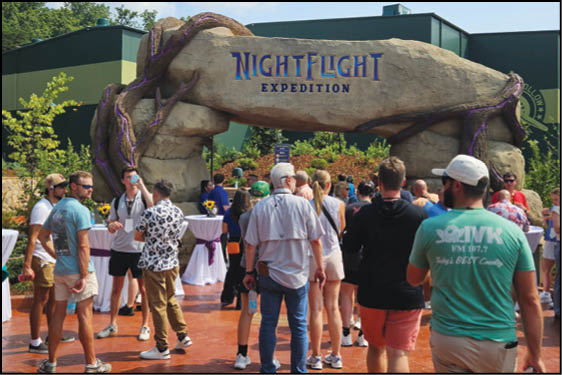 A group of people wearing casual summer clothing are gathered outdoors near a large stone archway with the words Night Flight Expedition written on it. Description generated by AI