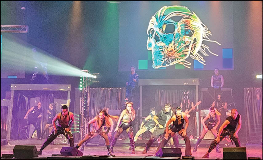 A group of people dressed in coordinated performance outfits are dancing energetically on a stage with colorful lighting and a large digital skull graphic projected in the background. Description generated by AI
