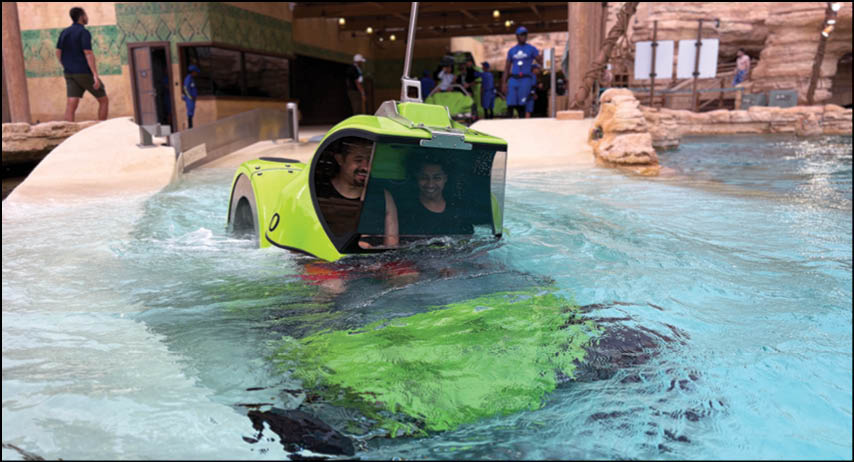 Two adults are riding in a small green amphibious vehicle partially submerged in a pool with people and a rocky structure in the background. Description generated by AI