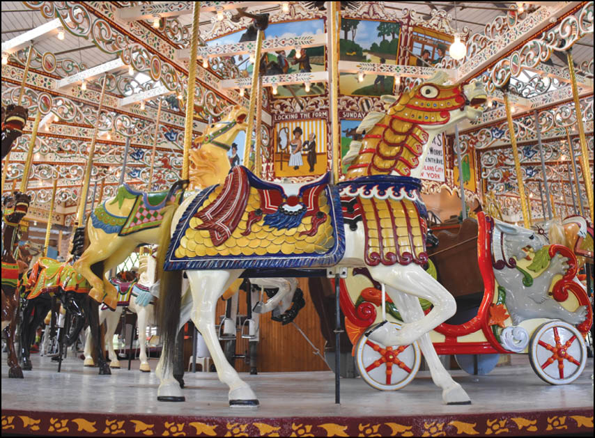 A colorful carousel with intricately decorated horses and a chariot is set indoors with bright lights and detailed artwork on the ceiling. Description generated by AI