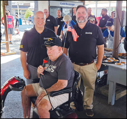 Three adults stand and sit together at an outdoor event with one person seated on a mobility scooter holding a microphone and a small American flag attached to the scooter. Description generated by AI