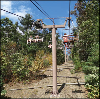 People are riding a chairlift above a wooded hillside on a clear, sunny day. Description generated by AI