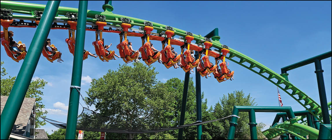 A group of people wearing orange clothing are riding a green and orange inverted roller coaster against a clear blue sky with trees in the background. Description generated by AI