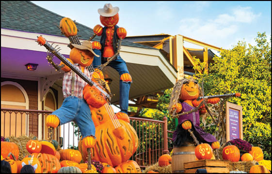 Three pumpkin-headed figures dressed in colorful clothing are playing musical instruments among a pile of pumpkins in an outdoor autumn setting. Description generated by AI