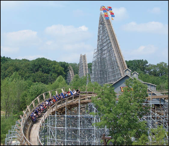 A group of people is riding a wooden roller coaster surrounded by trees with a tall, steep section of the coaster visible in the background under a partly cloudy sky. Description generated by AI