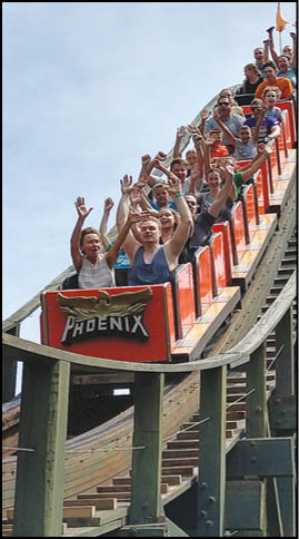 A group of people with raised hands are riding a wooden roller coaster named Phoenix on a clear day. Description generated by AI