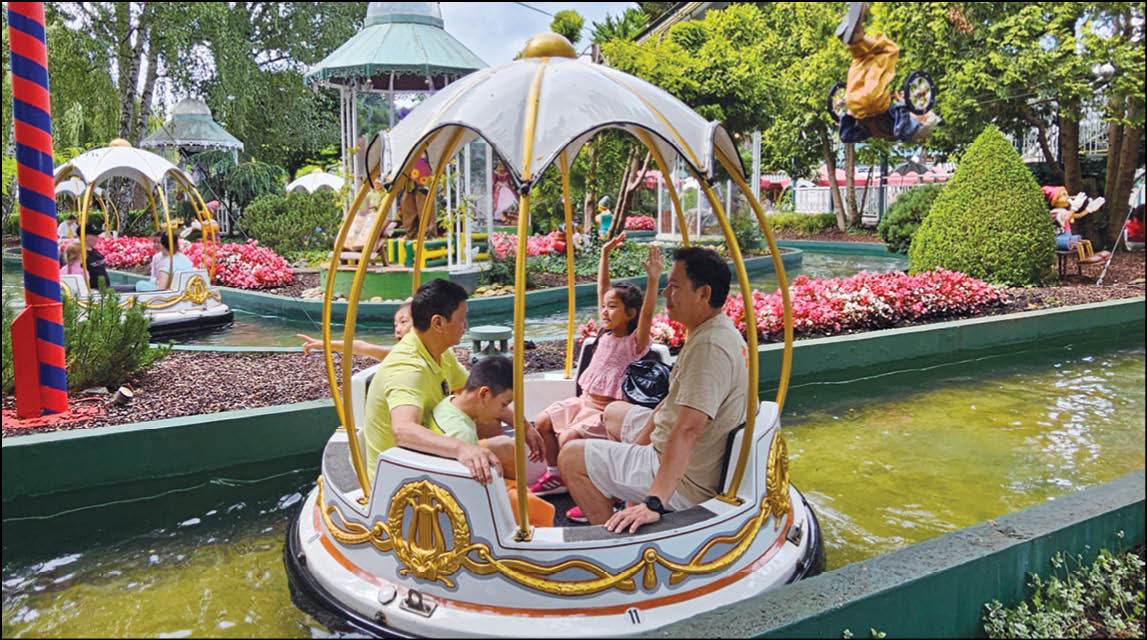 Four people are seated inside a decorative, round, canopy-covered boat floating on a narrow water channel in a park with greenery and flowers. Description generated by AI