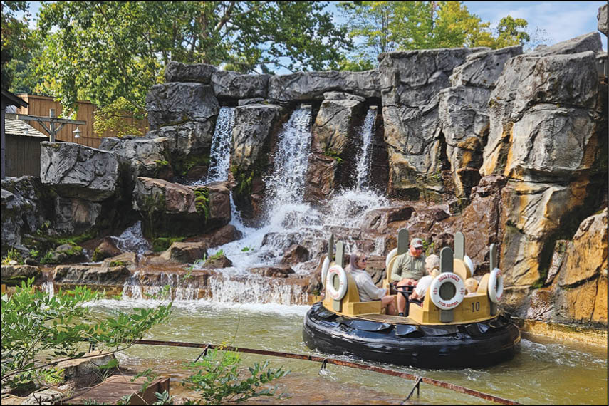Three people wearing casual clothing are riding a circular raft on a water ride with a rocky waterfall backdrop surrounded by greenery. Description generated by AI