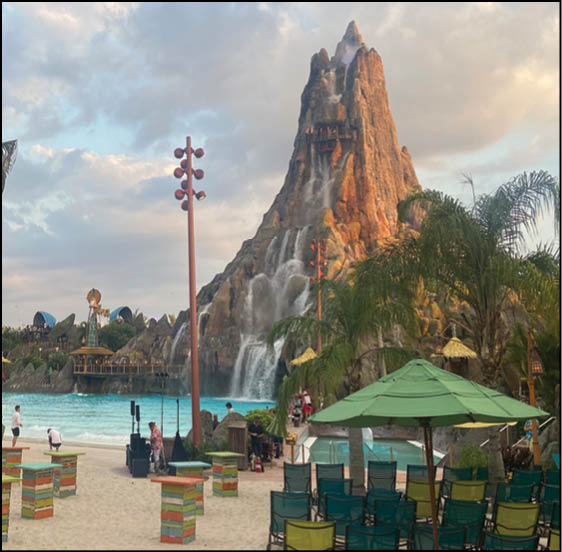 A large artificial volcano with cascading waterfalls stands behind a sandy beach area with colorful tables, chairs, umbrellas, and people near a pool and water attraction. Description generated by AI