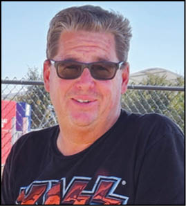 A person wearing sunglasses and a black graphic t-shirt is smiling outdoors in front of a chain-link fence on a sunny day. Description generated by AI