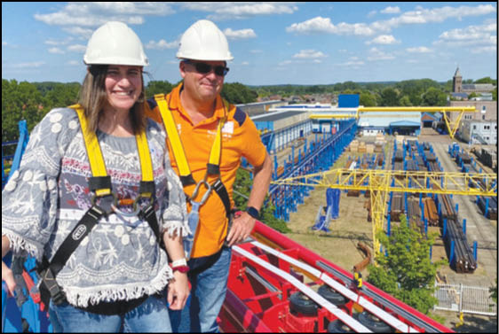 Two adults wearing white safety helmets and harnesses stand smiling at an industrial site with blue metal structures and yellow cranes under a partly cloudy sky. Description generated by AI