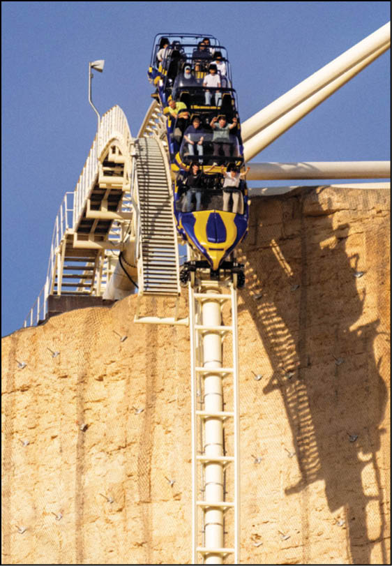 A roller coaster car filled with people is descending a steep track next to a rocky cliff under a clear blue sky. Description generated by AI
