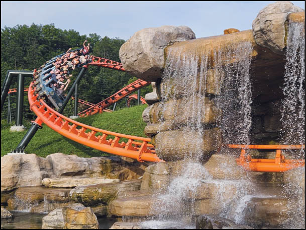 Adults are riding a roller coaster with orange tracks that curves around a large rock formation with a waterfall. Description generated by AI