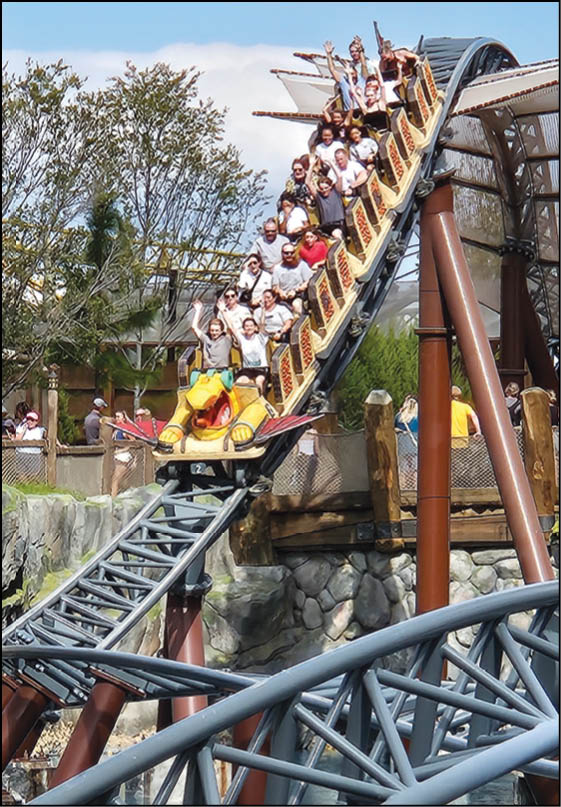 A group of people wearing casual clothing are riding a roller coaster descending a steep track outdoors with trees and a stone wall in the background. Description generated by AI