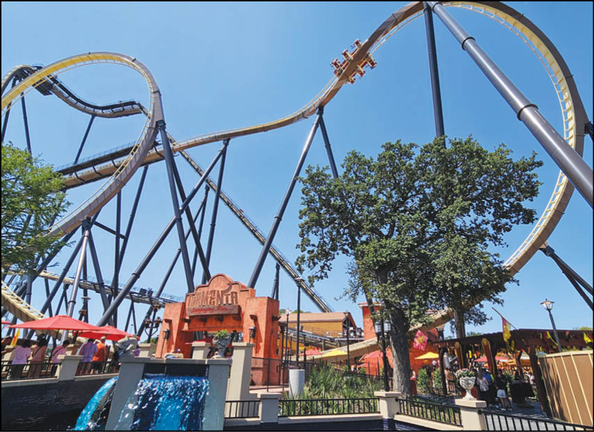 A large roller coaster with yellow tracks and black supports towers over a theme park area with trees, umbrellas, and people walking around. Description generated by AI