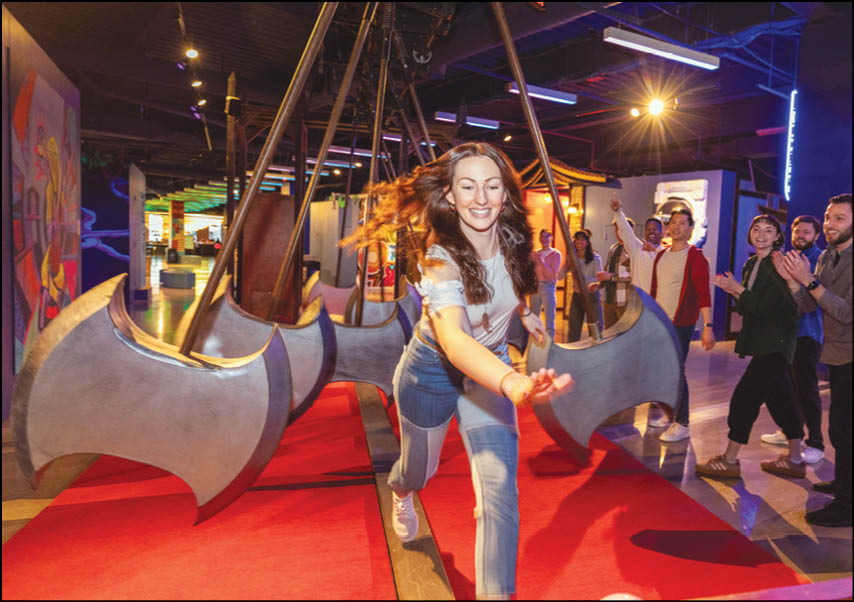 A person with long hair wearing a white shirt and blue jeans is running excitedly on a red carpet in an indoor setting with large swinging axe-shaped objects and several people watching in the background. Description generated by AI