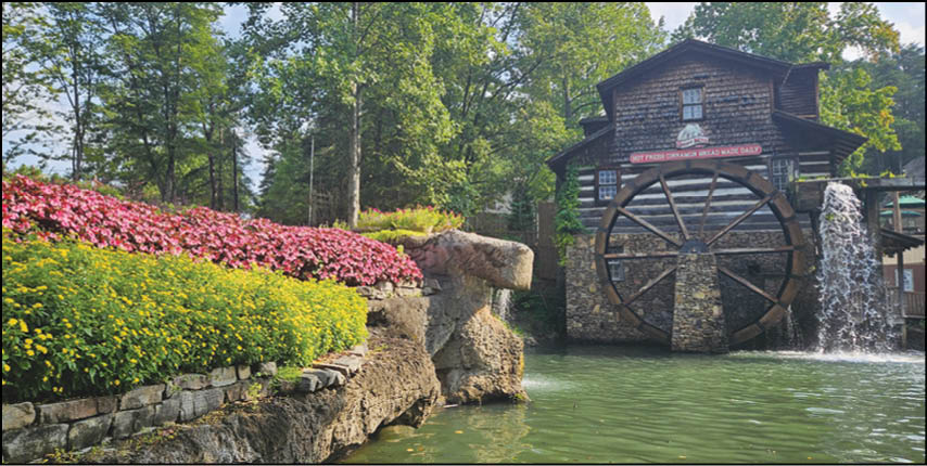 A rustic watermill with a large wooden wheel is situated beside a pond surrounded by vibrant yellow and red flowers and lush green trees. Description generated by AI