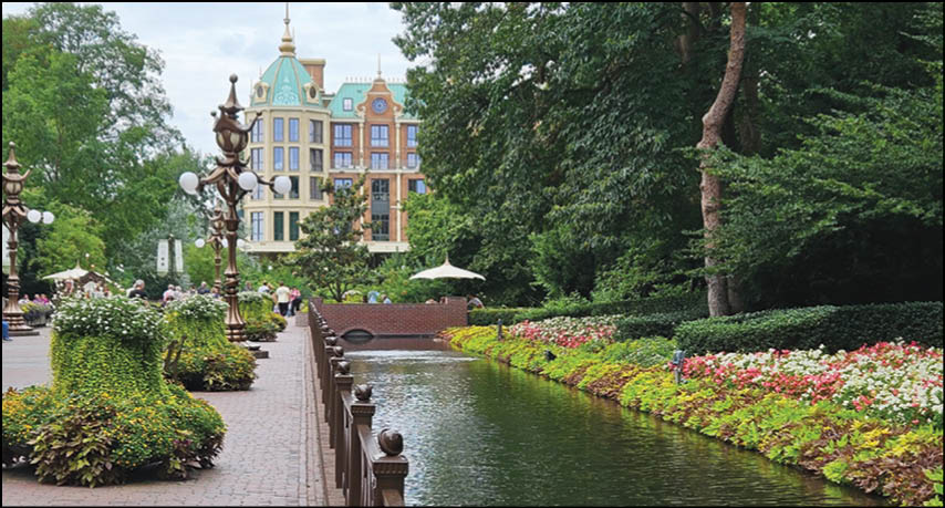 A serene park scene with a narrow water canal bordered by colorful flowers and greenery, a paved walkway with benches and vintage-style street lamps, and a large ornate building in the background. Description generated by AI