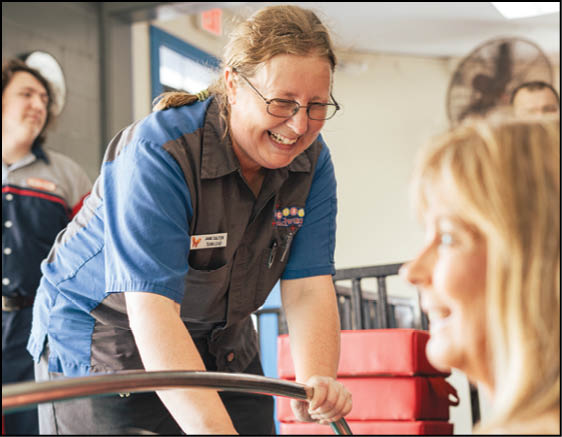 A person wearing glasses and a blue and gray uniform is smiling and leaning forward while interacting with another person who is slightly out of focus in the foreground. Description generated by AI