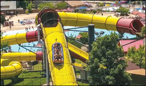 People are riding a yellow and red enclosed water slide at a water park surrounded by trees and other attractions. Description generated by AI