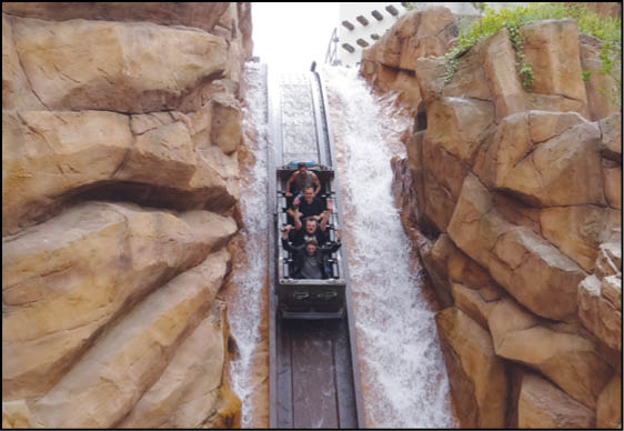 Four people are riding a log flume water ride descending between large artificial rock formations with water splashing on both sides. Description generated by AI
