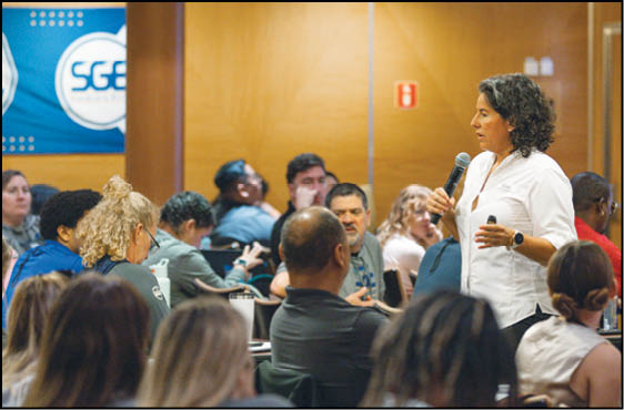 An adult with curly hair wearing a white shirt is speaking into a microphone while standing in front of a seated audience in a conference room. Description generated by AI