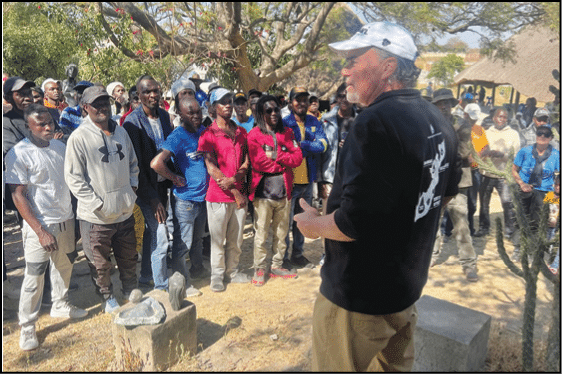 A person wearing a black shirt and white cap is speaking to a large group of people standing outdoors near trees and stone blocks. Description generated by AI
