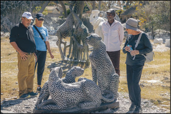 Five people stand outdoors around a stone sculpture of three large spotted cats with a giraffe sculpture in the background. Description generated by AI