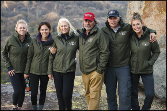 Six adults wearing matching green jackets stand closely together outdoors on rocky terrain with a natural landscape in the background. Description generated by AI