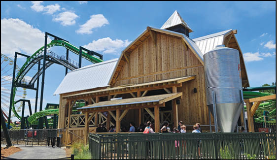 A wooden building with a metal roof is next to a green roller coaster track with people standing near the building under a clear blue sky. Description generated by AI