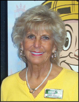 A person with short, styled hair wearing a yellow top and a name badge is smiling in front of a colorful cartoon background. Description generated by AI
