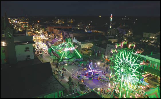 A nighttime aerial view of a brightly lit carnival with illuminated rides including a Ferris wheel and spinning attractions surrounded by people and buildings. Description generated by AI