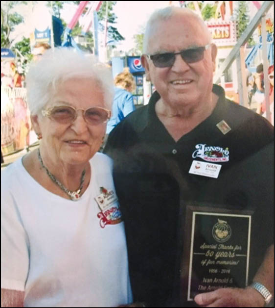 Two seniors wearing glasses and event name tags smile while one holds a plaque at an outdoor fair or carnival. Description generated by AI