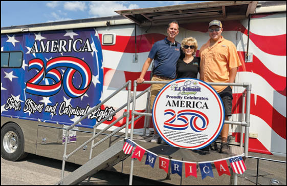 Three adults stand smiling on a metal ramp in front of a decorated vehicle with patriotic red, white, and blue colors and signs celebrating America 250. Description generated by AI