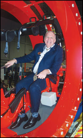 A person in a suit and tie is sitting and smiling inside a large red circular structure with harnesses and mechanical components. Description generated by AI