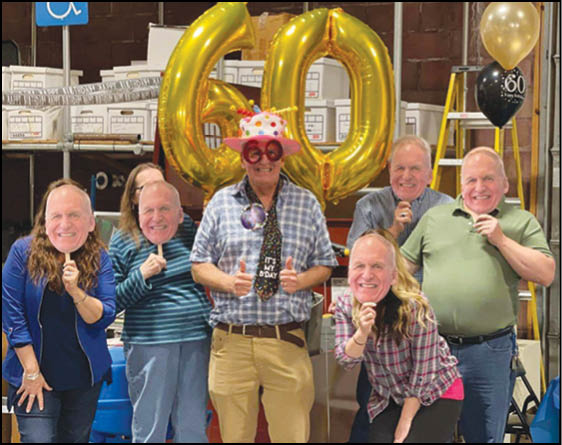 A group of six people wearing large smiling face masks pose cheerfully in front of gold balloons shaped like the number 60 and festive decorations. Description generated by AI